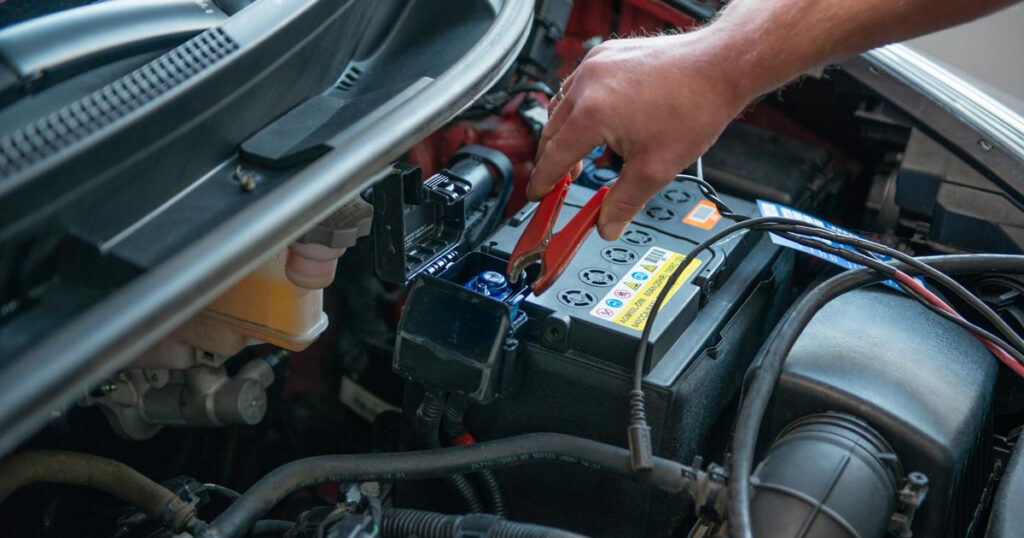 Mechanikerhand setzt ein Starthilfekabel an die Autobatterie im Motorraum an.