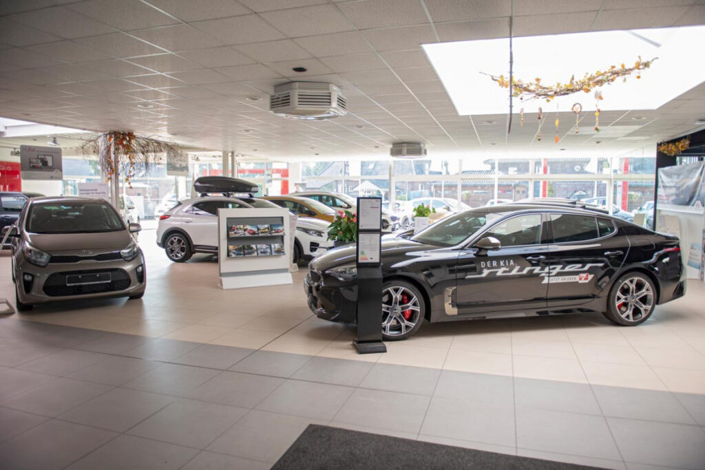 Heller Autohaus-Showroom mit mehreren ausgestellten Kia-Fahrzeugen, darunter ein schwarzer Kia Stinger und mehrere kompakte Modelle, präsentiert zwischen großen Fenstern und Displayständern