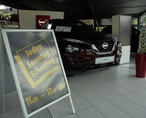 Verkaufsraum mit einem Hinweisschild Jeden Sonntag Schautag 11 bis 17 Uhr mi einem schwarzen Nissan im Hintergrund.
