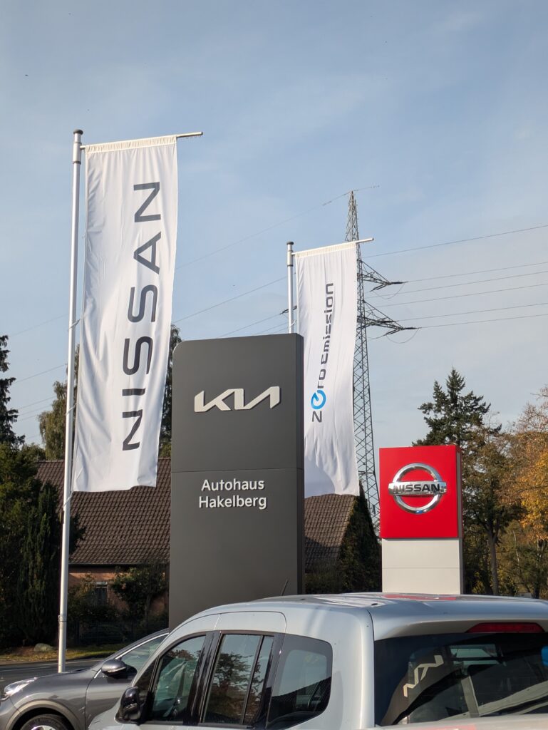 Eingangsbereich des Autohaus Hakelberg mit KIA- und Nissan-Logos sowie Fahnen vor blauem Himmel.
