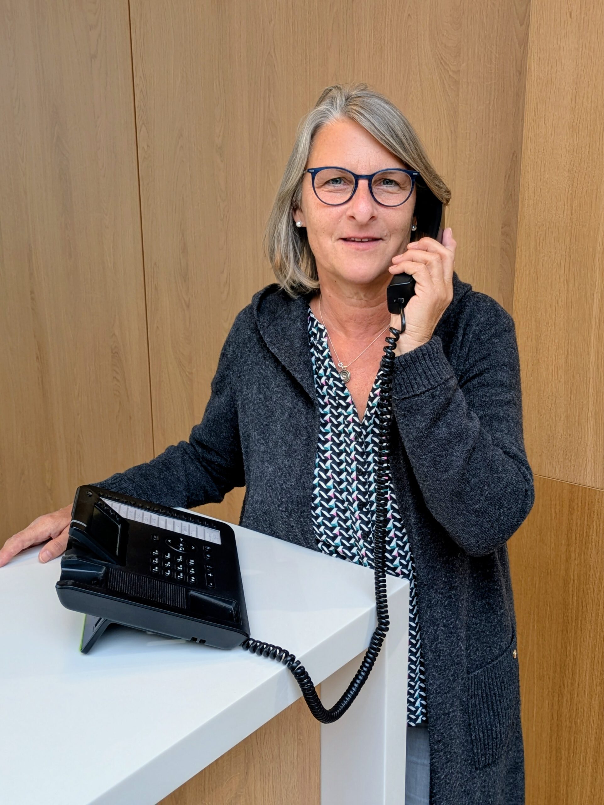 Mitarbeiterin im KIA-Servicebereich steht lächelnd an einem weißen Tresen vor Holzrückwand bei einem Kundentelefonat.