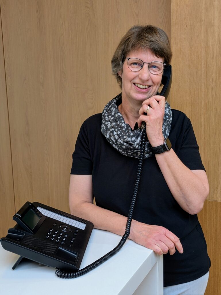 Mitarbeiterin mit Brille im KIA-Servicebereich steht lächelnd an einem weißen Tresen vor Holzrückwand bei einem