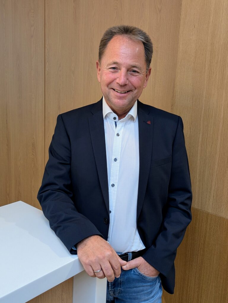 Mitarbeiter Herr Schwarze im KIA-Servicebereich steht lächelnd an einem weißen Tresen vor Holzrückwand.