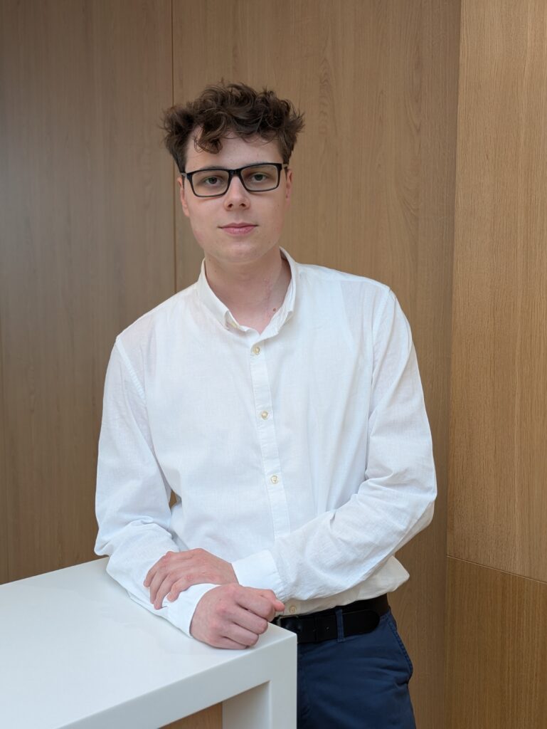 Mitarbeiter Niklas Ude im KIA-Servicebereich steht lächelnd an einem weißen Tresen vor Holzrückwand.